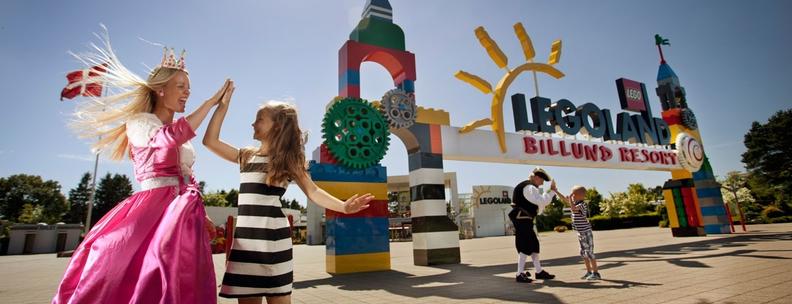 Mujer vestida con un disfraz de princesa rosa, chocando los cinco con una niña con un vestido a rayas negras y blancas, frente a la entrada del parque de atracciones Legoland
